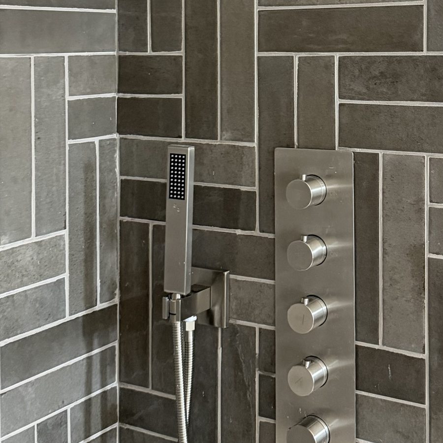 Thermostatic shower control stack with four handles and a handheld sprayer on a herringbone tiled wall beside a recessed pebble niche