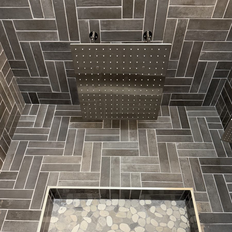 Looking down at a large square rain shower panel mounted overhead, with herringbone tile walls and a pebble-filled pan below