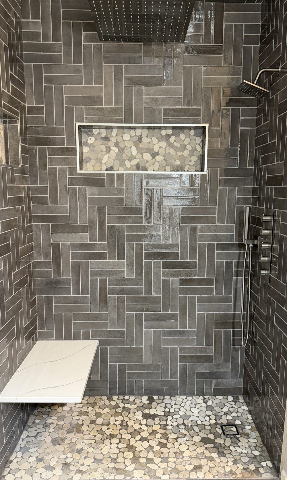 Finished walk-in shower tiled in dark grey herringbone from floor to ceiling, with a recessed pebble niche, a floating white bench, a thermostatic control stack and a pebble stone floor