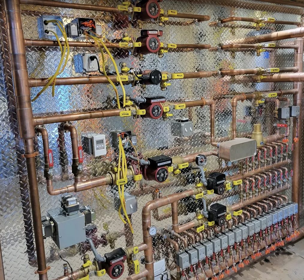 A copper radiant-heat manifold with brass valves and circulator pumps mounted on a wall, built by Top Tier