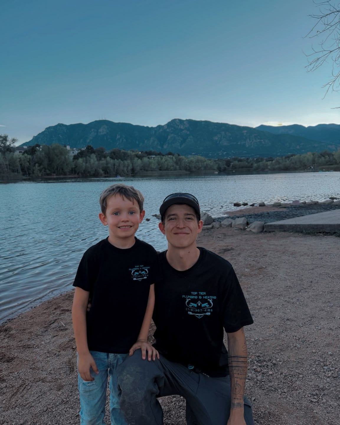 Tate of Top Tier Plumbing & Heating kneeling lakeside with his son, both in matching Top Tier shirts, the front range behind them