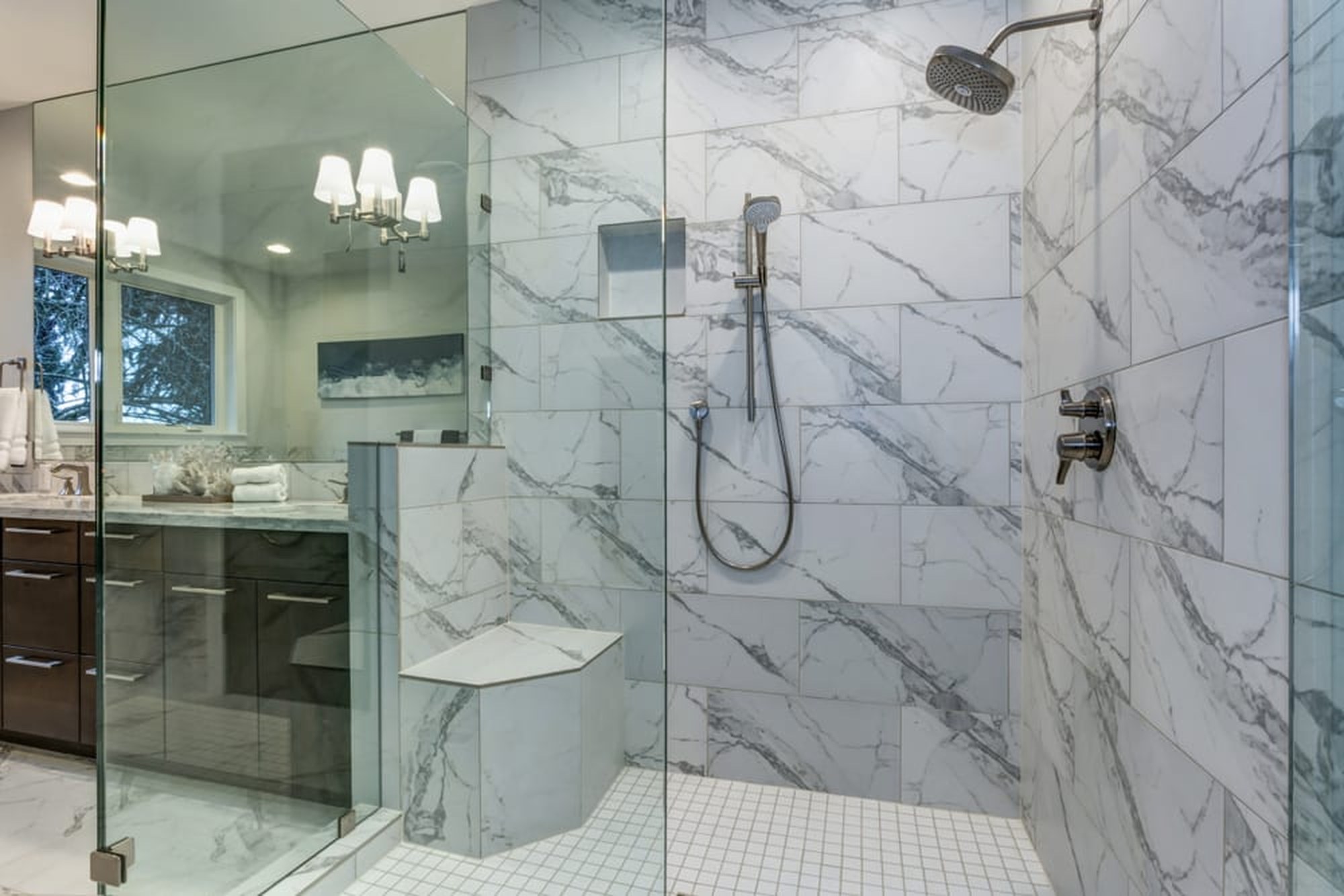 A marble-tiled walk-in shower with a glass enclosure, rain head, handheld sprayer on a slide bar and a corner bench, beside a dark wood vanity