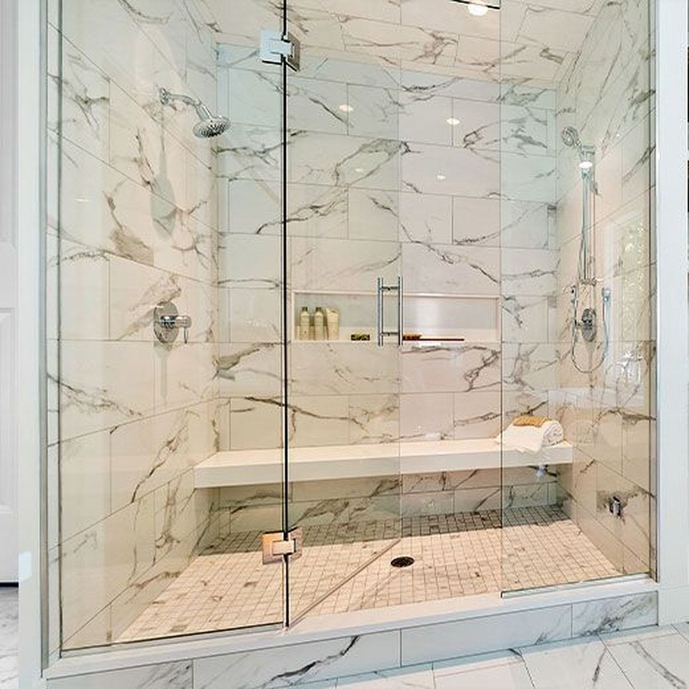 A wide double shower in marble tile with two shower heads, two valve sets, a full-length bench and a recessed niche